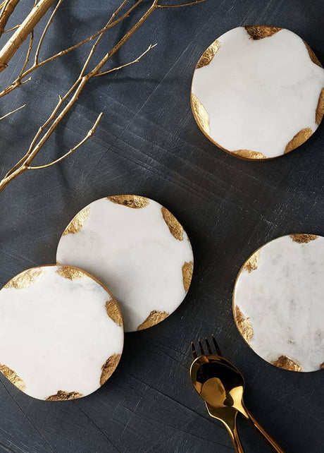 Round white marble coasters with gold leaf accents on a dark surface, styled with gold branches and cutlery.