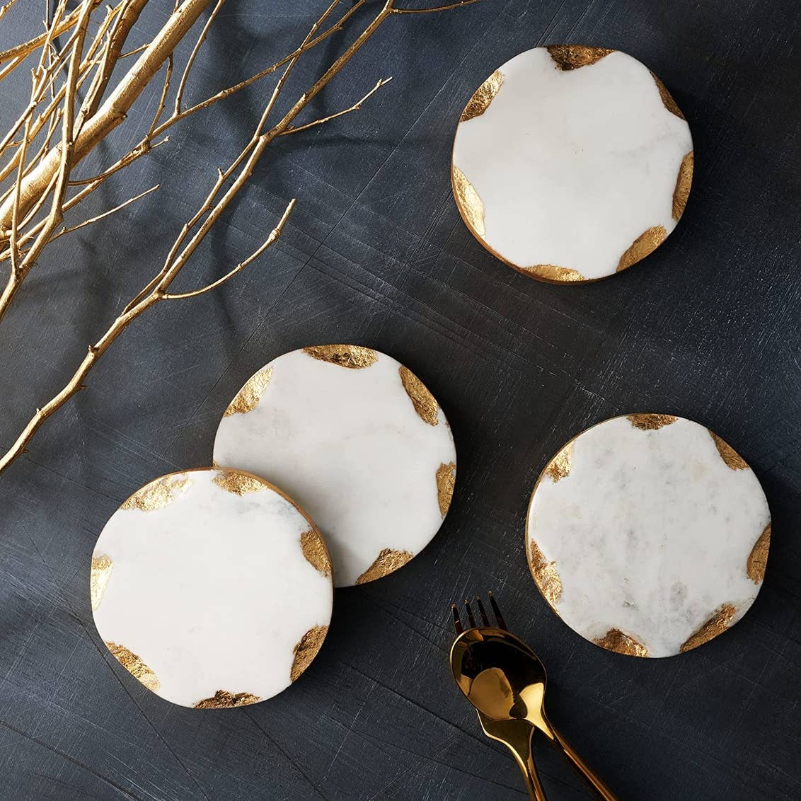Round white marble coasters with gold leaf accents on a dark surface, styled with gold branches and cutlery.
