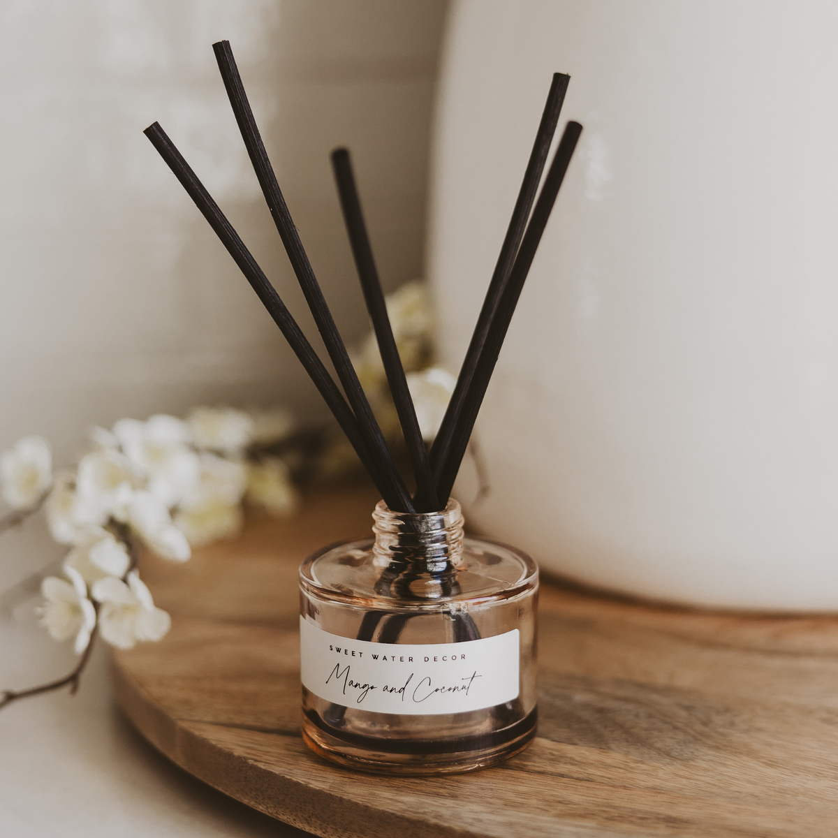 Mango and Coconut reed diffuser on a wooden tray with white flowers, bringing warmth and beauty to everyday living.